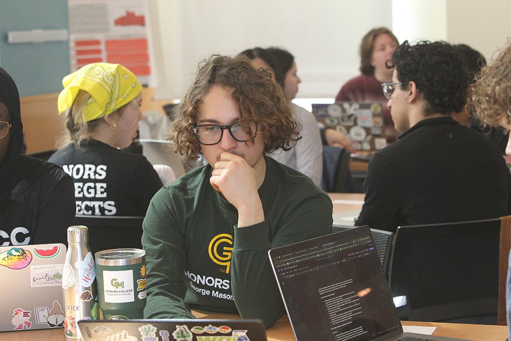 A student looking thoughtfully at their laptop, surrounded by students with laptops open working in teams.